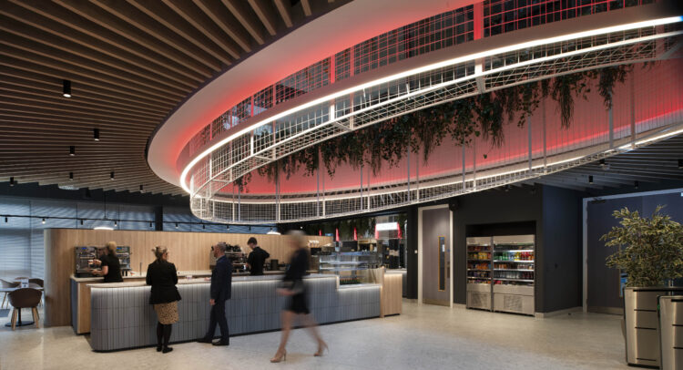 An expansive office reception and cafe area, people roam the central atrium space as wooden panels expand outwards from a central glass atrium that cuts through the entire office building.