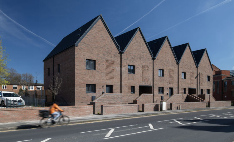 View of 1 red brick terrace of 5 homes fronting a road and with a blue sky in the background. A cyclist passes by the front. The Parkmount development pictured is a passivhaus project.