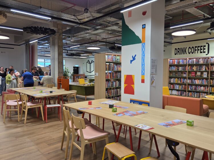 A modern community library and social space in Stockport, Stockroom designed by AEW Architects, featuring an inviting interior with a blend of open seating, bookshelves, and colourful decor. The walls are adorned with bold, abstract art and signage like "DRINK COFFEE," adding a playful, welcoming atmosphere. The space has wooden tables and chairs in soft tones of pink and yellow, fostering an informal and inclusive environment. People are browsing books, chatting, and queuing at the counter, creating a lively, communal scene. Exposed industrial ceilings with lighting fixtures and natural timber finishes further contribute to the contemporary design.