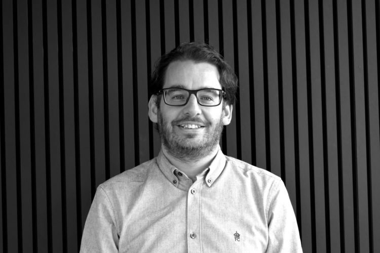 A black and white portrait of a man, AEW Architects Director Mark Turner, with dark hair, wearing glasses and a shirt. He is smiling and standing in front of a vertical slatted background.