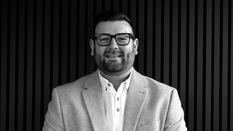 Associate Architect and Retrofit Coordinator Danny Lomax from AEW Architects wearing a white shirt and a gray blazer against a dark backdrop.