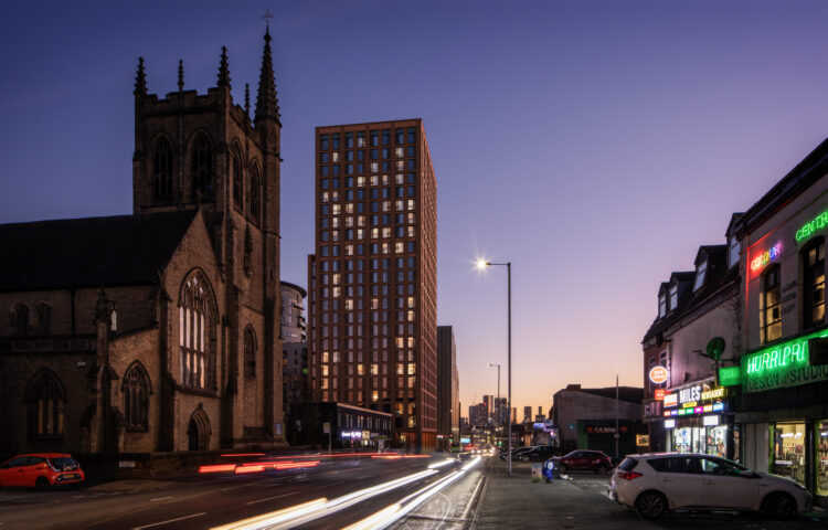 A 24 storey residential tower designed by AEW Architects on Lord Street, Manchester. This view shows the development at dusk with the building against a purple sky. In the foreground is a church and the lights of moving traffic.